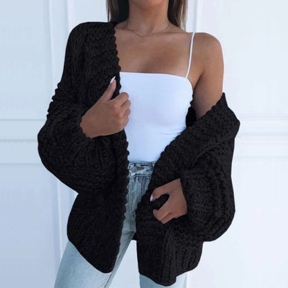 Oversize-Grobstrickjacke in Schwarz, offen über einem weißen Top getragen, vor einer weißen Kassettenwand
