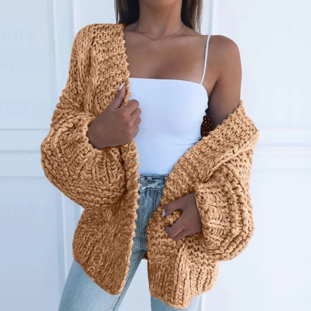 Oversize-Grobstrickjacke in Camel, offen über einem weißen Top getragen, vor einer weißen Kassettenwand