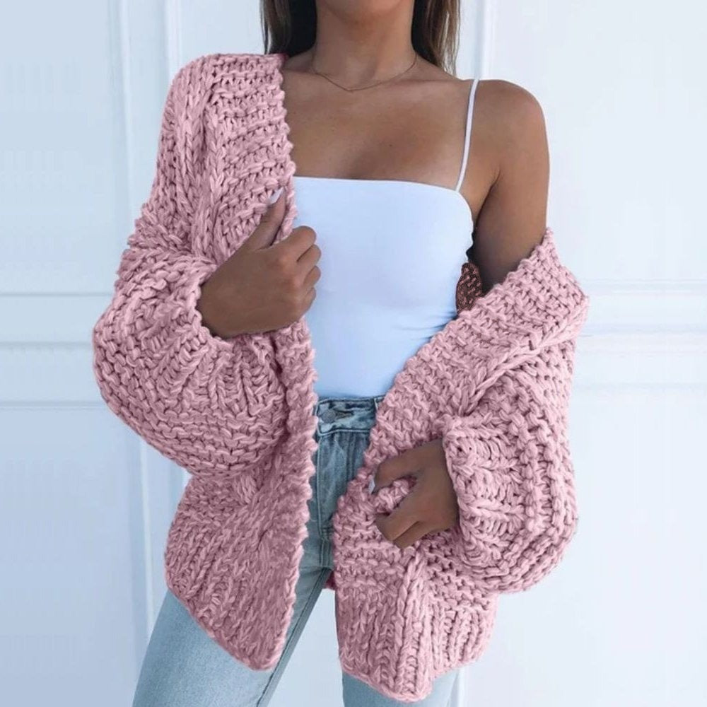 Oversize-Grobstrickjacke in Rosa, offen über einem weißen Top getragen, vor einer weißen Kassettenwand