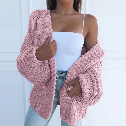 Oversize-Grobstrickjacke in Rosa, offen über einem weißen Top getragen, vor einer weißen Kassettenwand