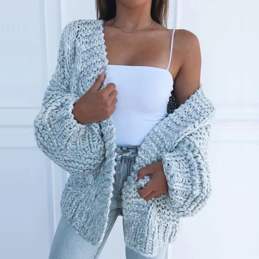 Oversize-Grobstrickjacke in Hellblau, offen über einem weißen Top getragen, vor einer weißen Kassettenwand